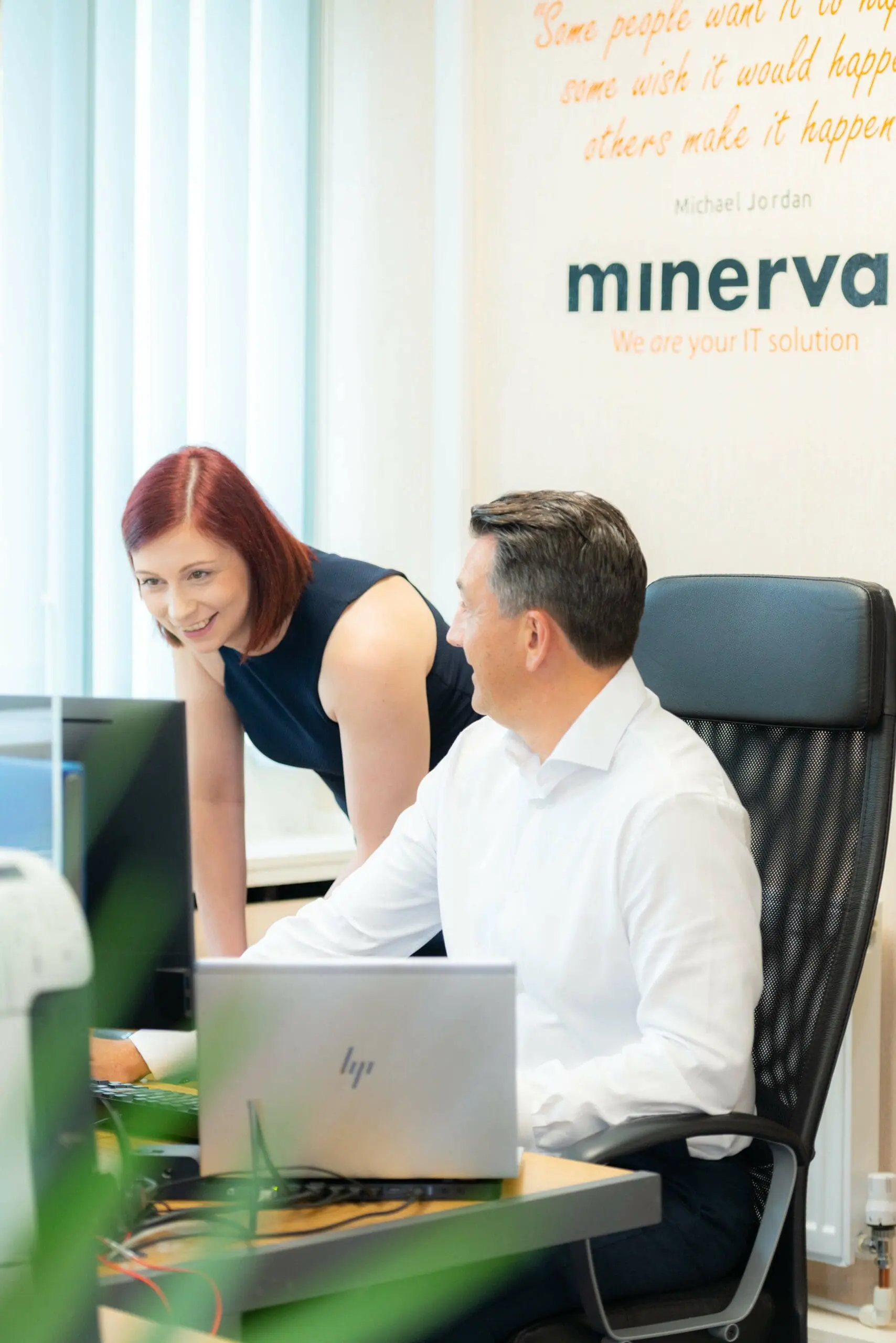 a woman leans over a desk assisting a man seated in a black chair, both engaged in a discussion at a modern office workspace.