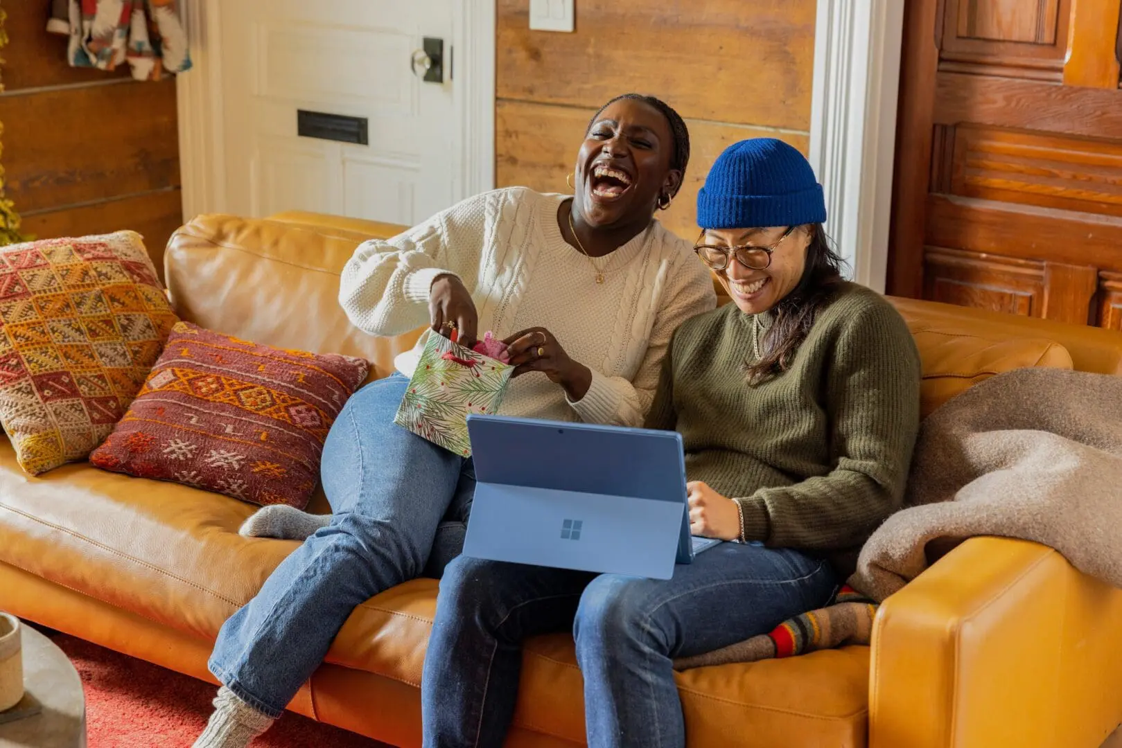 two people sit on a brown leather couch, one opening a gift while the other studies a tablet. colorful pillows and a cozy atmosphere enhance the scene.